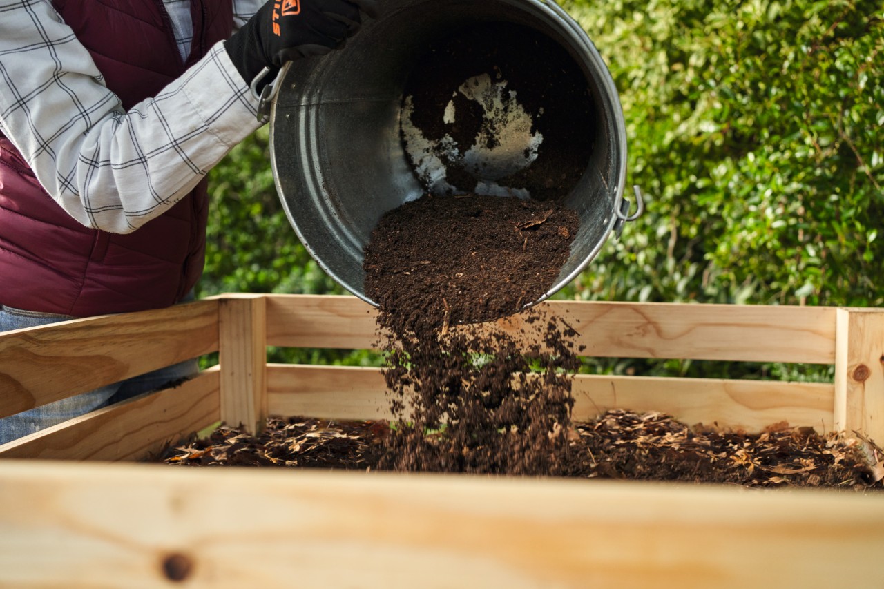 Eine Frau schüttet aus einem Eimer Gartenerde auf den Komposter
