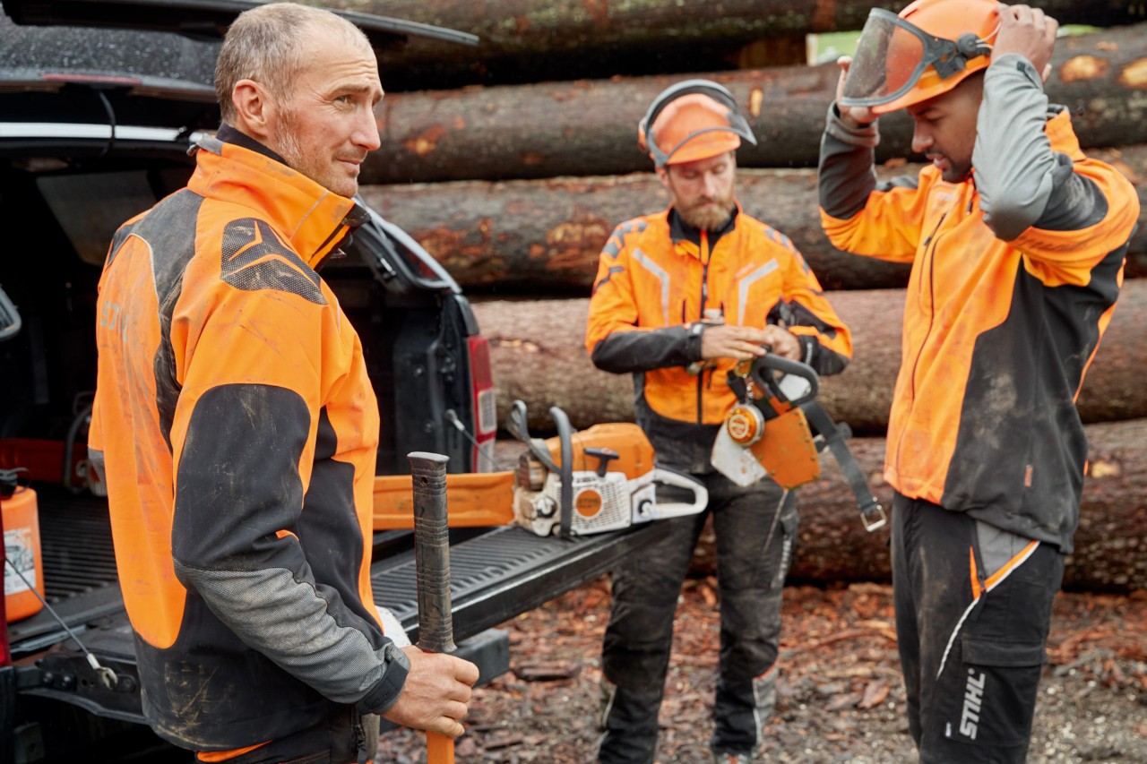 Троє чоловіків у захисному одязі STIHL перед штабелем деревини готуються до роботи з ланцюговою пилою STIHL