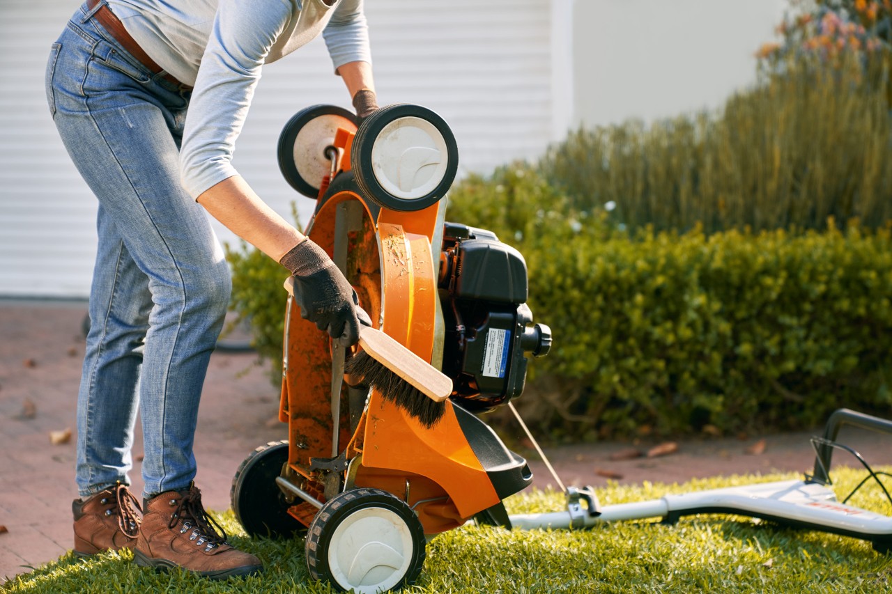 Frau reinigt STIHL Benzin-Rasenmäher RM 448 T-C mit einem Handfeger 