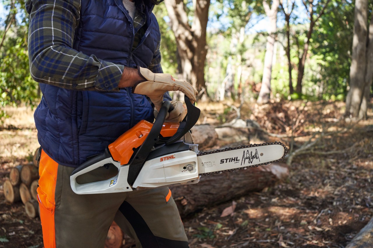 Чоловік у лісовій місцевості тримає бензинову мотопилу STIHL MS 162, на задньому плані стовбури дерев у штабелі