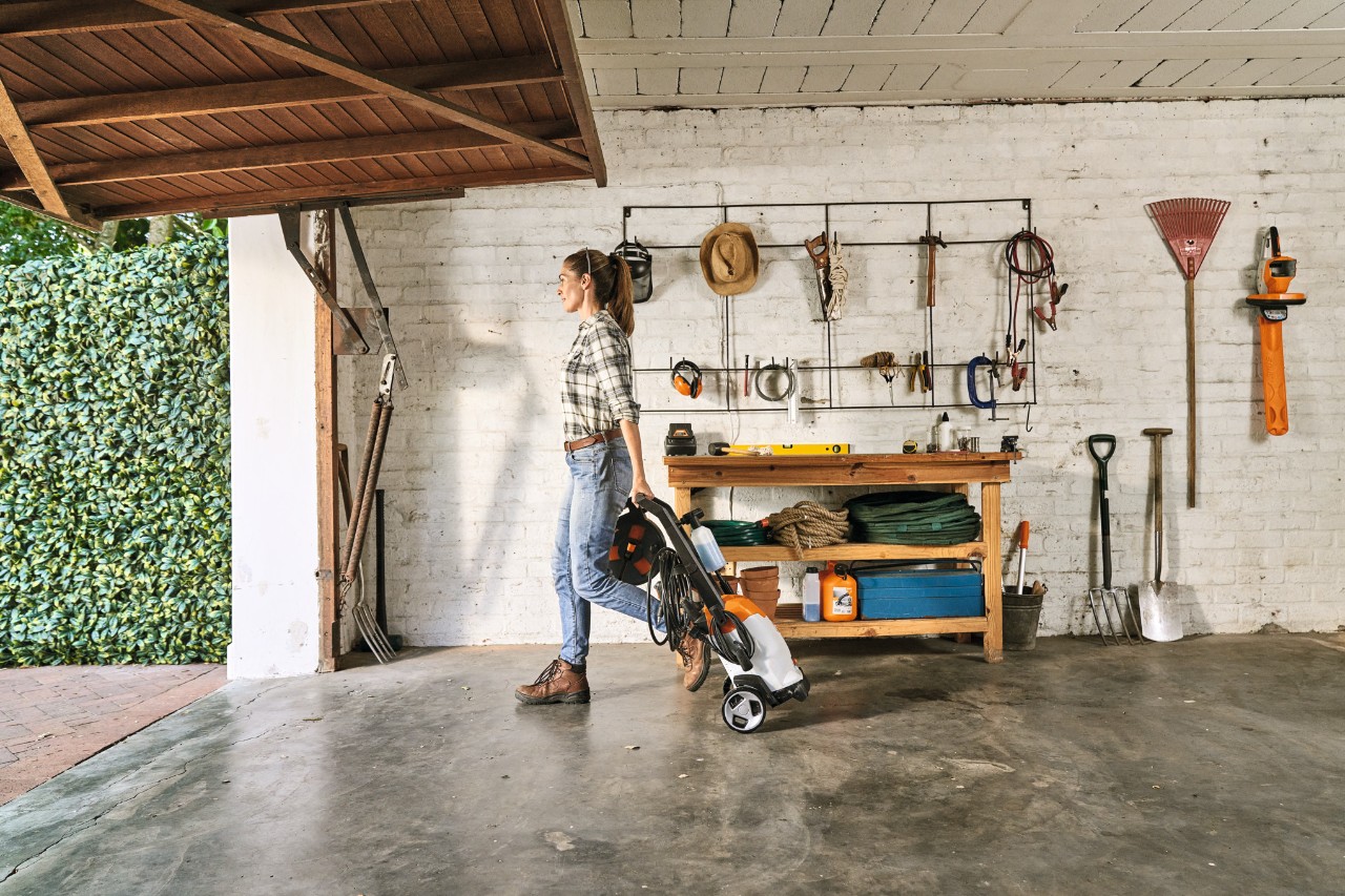 STIHL Felgenreiniger, Fahrzeugshampoo und Waschbürste auf dem Boden vor weißer Garage. Im Hintergrund ein STIHL Hochdruckreiniger