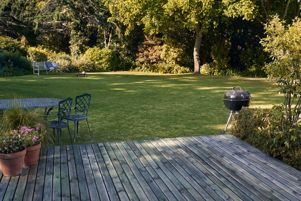 Holzterrasse vor einem grünen Garten mit Grill, Stühlen und Blumenkübeln
