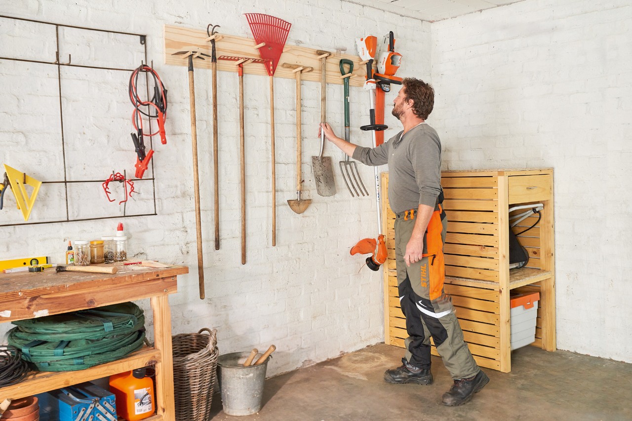 Mann in STIHL Arbeitshose hängt Schaufel an den selber gebauten Gartengerätehalter neben einem STIHL Rasentrimmer 