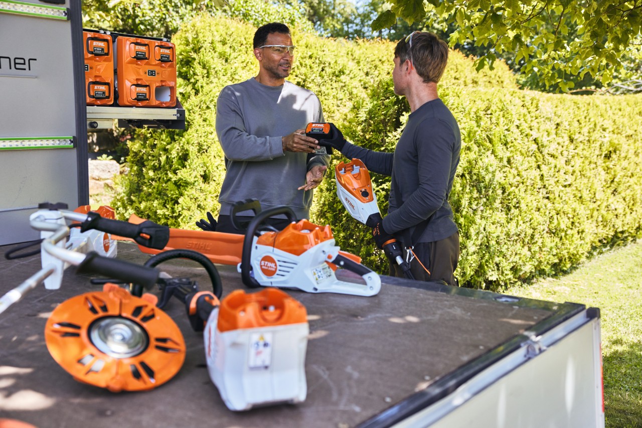 Чоловік передає своєму колезі акумулятор STIHL, обидва знаходяться позаду вантажної зони транспортного засобу, також там знаходиться bottTainer і акумуляторна мотопила STIHL, а також інше обладнання