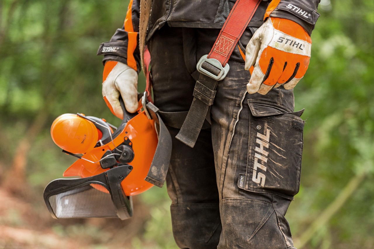 Людина в захисних штанях STIHL і рукавичках STIHL тримає в руці захисний шолом STIHL