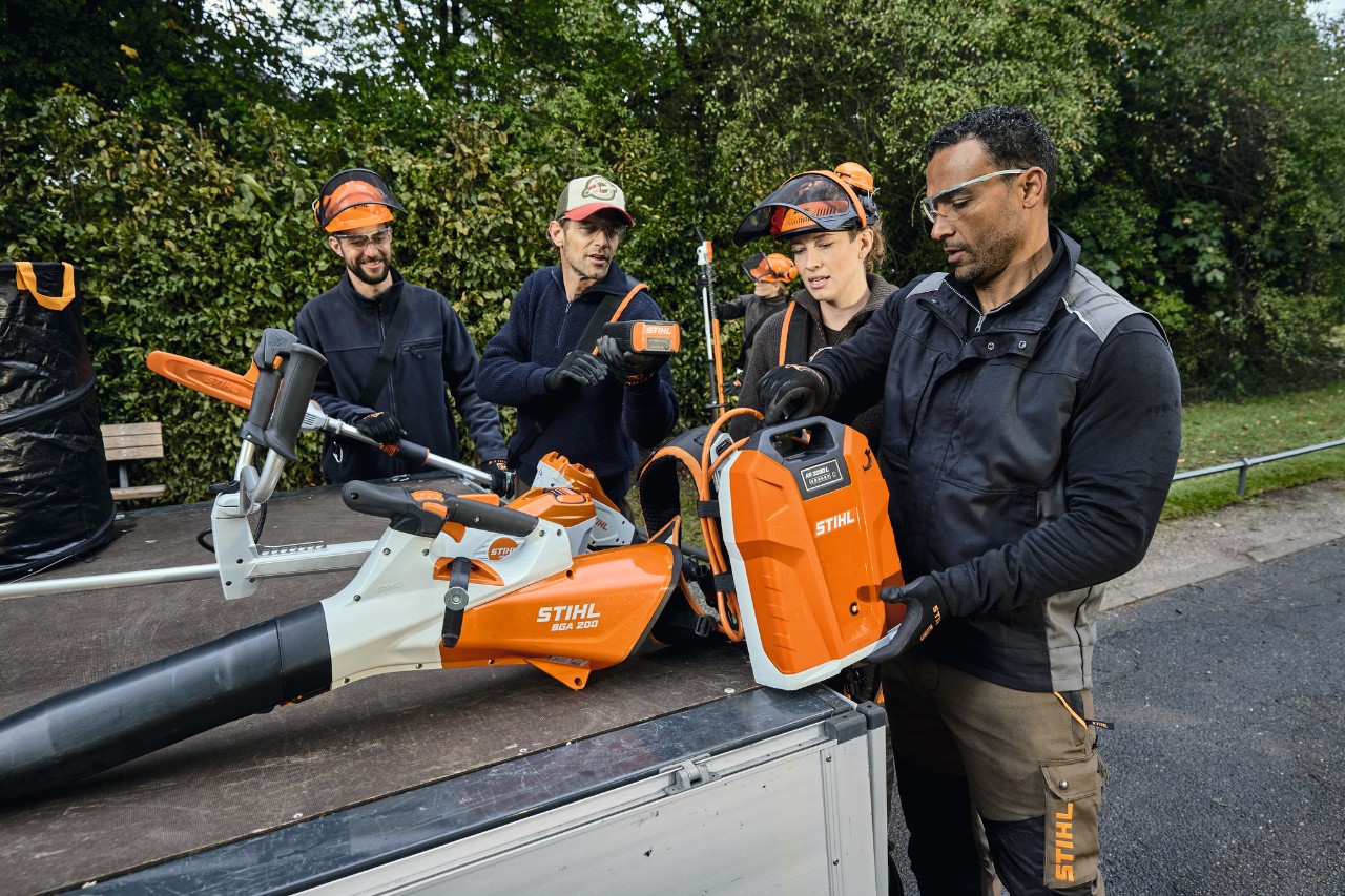 Троє чоловіків і жінка в захисному спорядженні STIHL стоять на вантажному майданчику автомобіля з інструментами STIHL, такими як акумуляторний повітродувний пристрій для листя STIHL BGA 200 і ранцевий акумулятор STIHL AR 2000 L