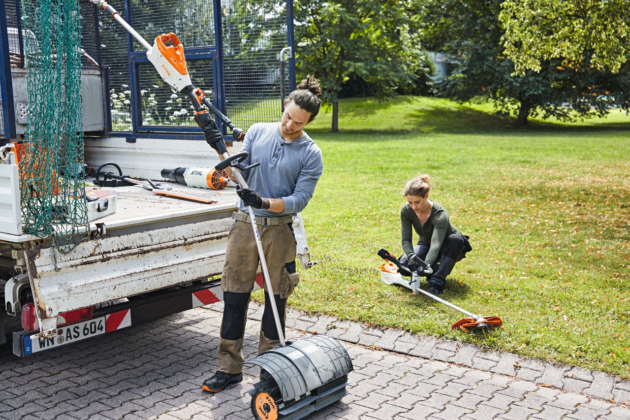 Чоловік і жінка працюють акумуляторними інструментами STIHL поряд із автомобілем із відкритою вантажною платформою, на задньому фоні луг