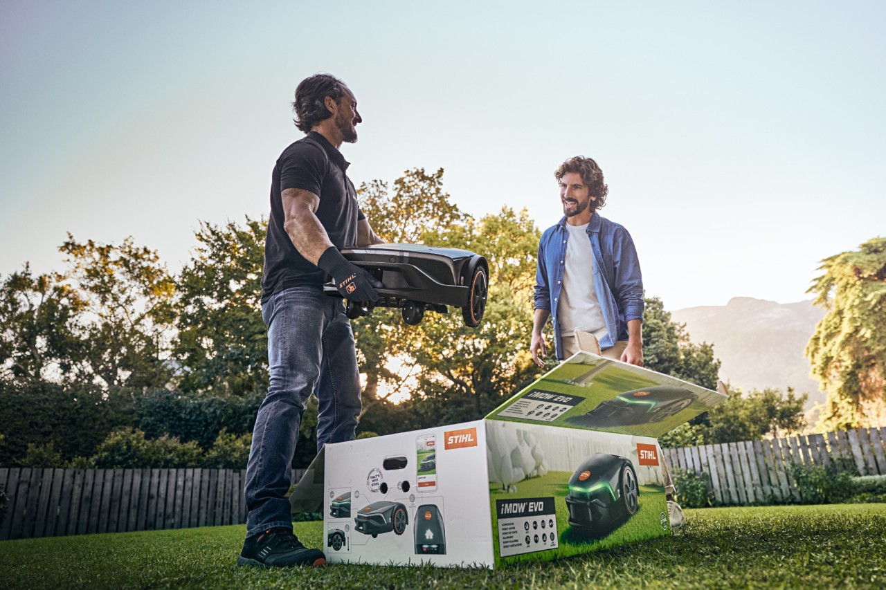 Спеціалізований дилер STIHL стоїть у саду клієнта з iMOW® EVO в руках і коробкою iMOW® перед ним і допомагає зі встановленням