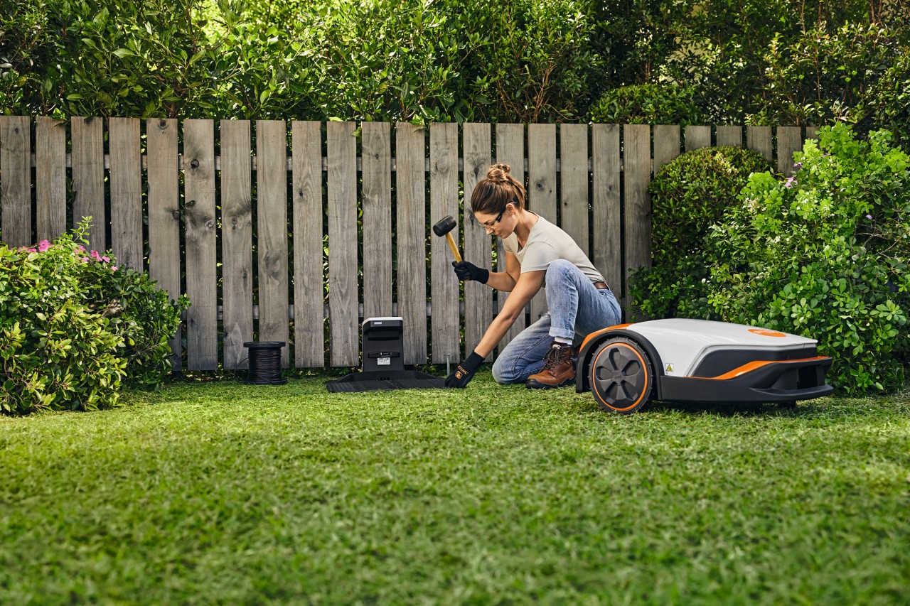 Жінка сама встановлює док-станцію для свого STIHL iMOW® перед парканом у своєму саду.